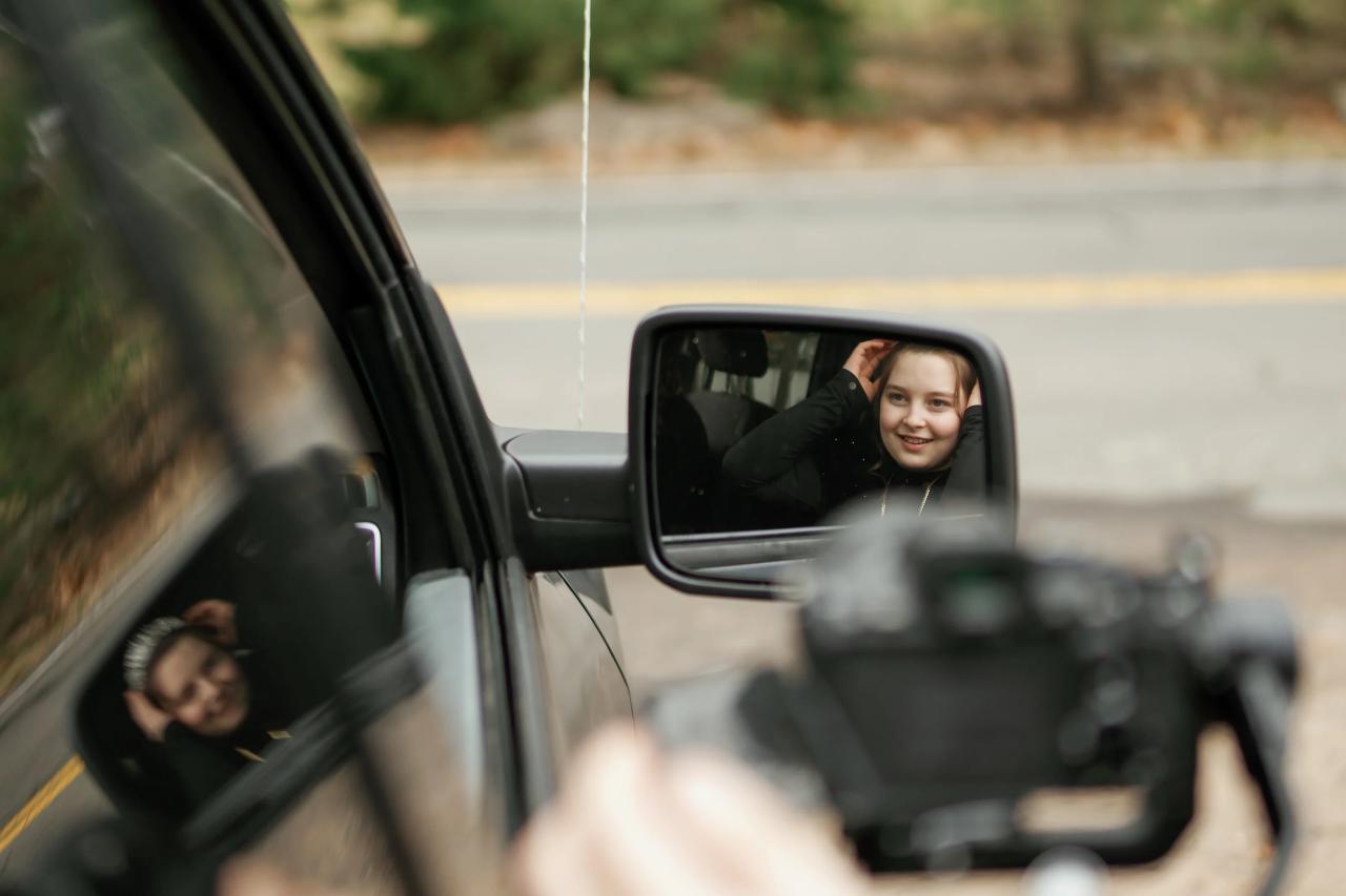 A candid outdoor moment framed through a car side mirror showing a smiling child in the back seat and a photographer’s camera in the foreground.