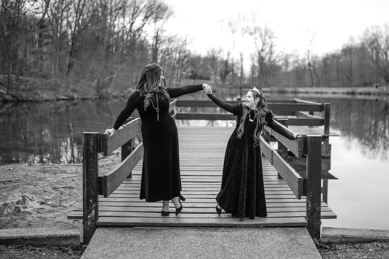 Black-and-white portrait of two women on a wooden boardwalk by a lake, walking together while holding out their arms in a playful, joyful moment.