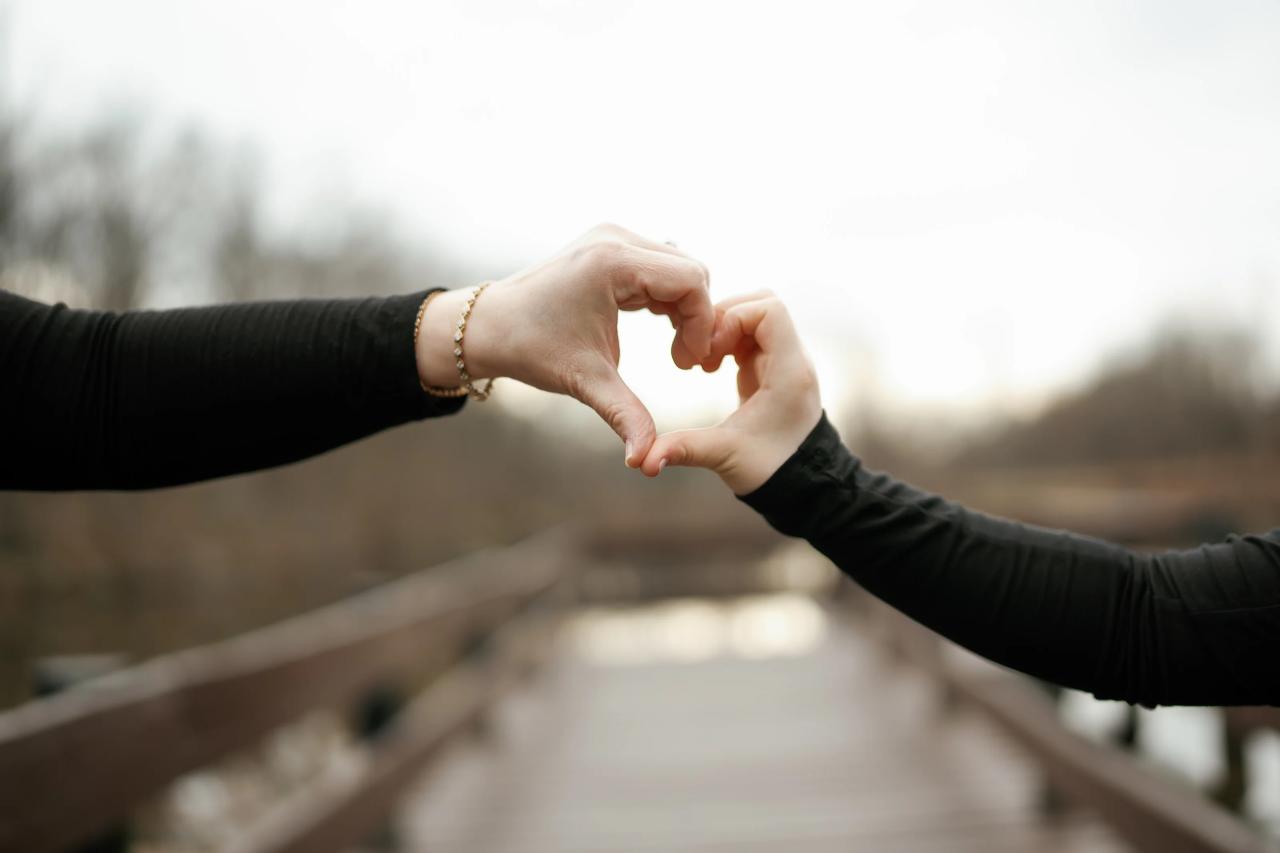 Close-up of two people’s hands forming a heart shape toward each other against a softly blurred outdoor background at golden hour.