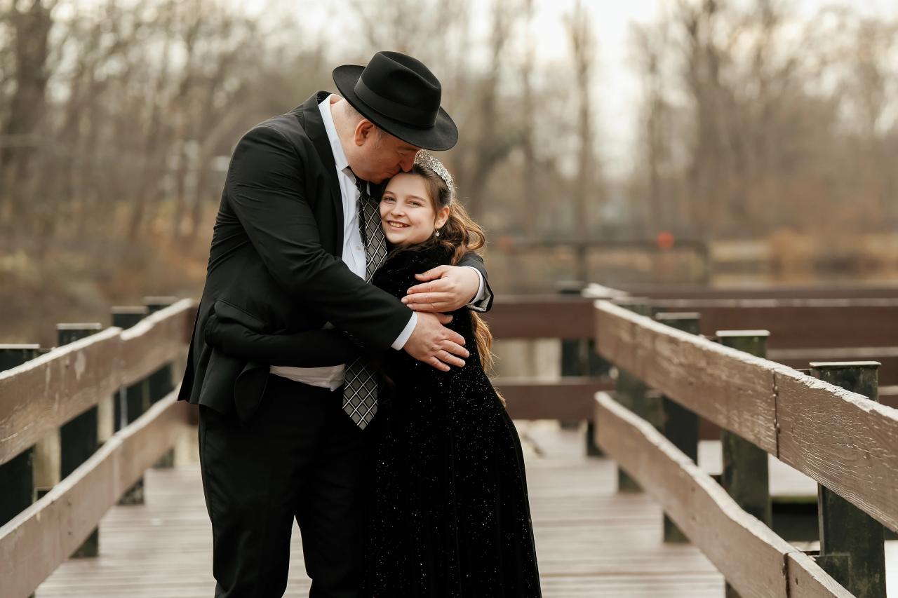 Man in a black hat embracing a young woman in an elegant dark outfit on an outdoor wooden walkway, both smiling with warmth.