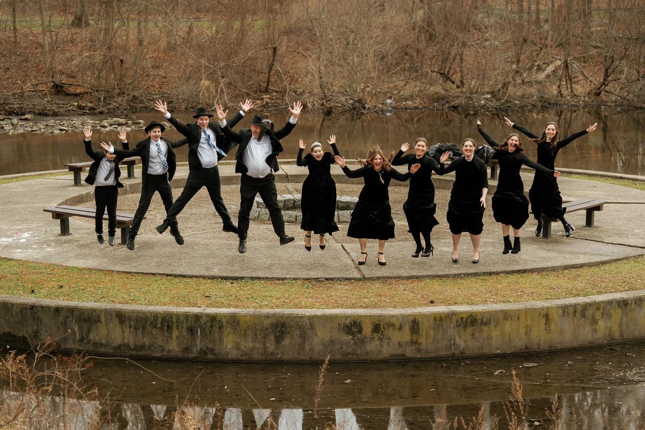 A group of guests in formal attire dancing outdoors with arms raised, smiling as they celebrate by a stone walkway and water.