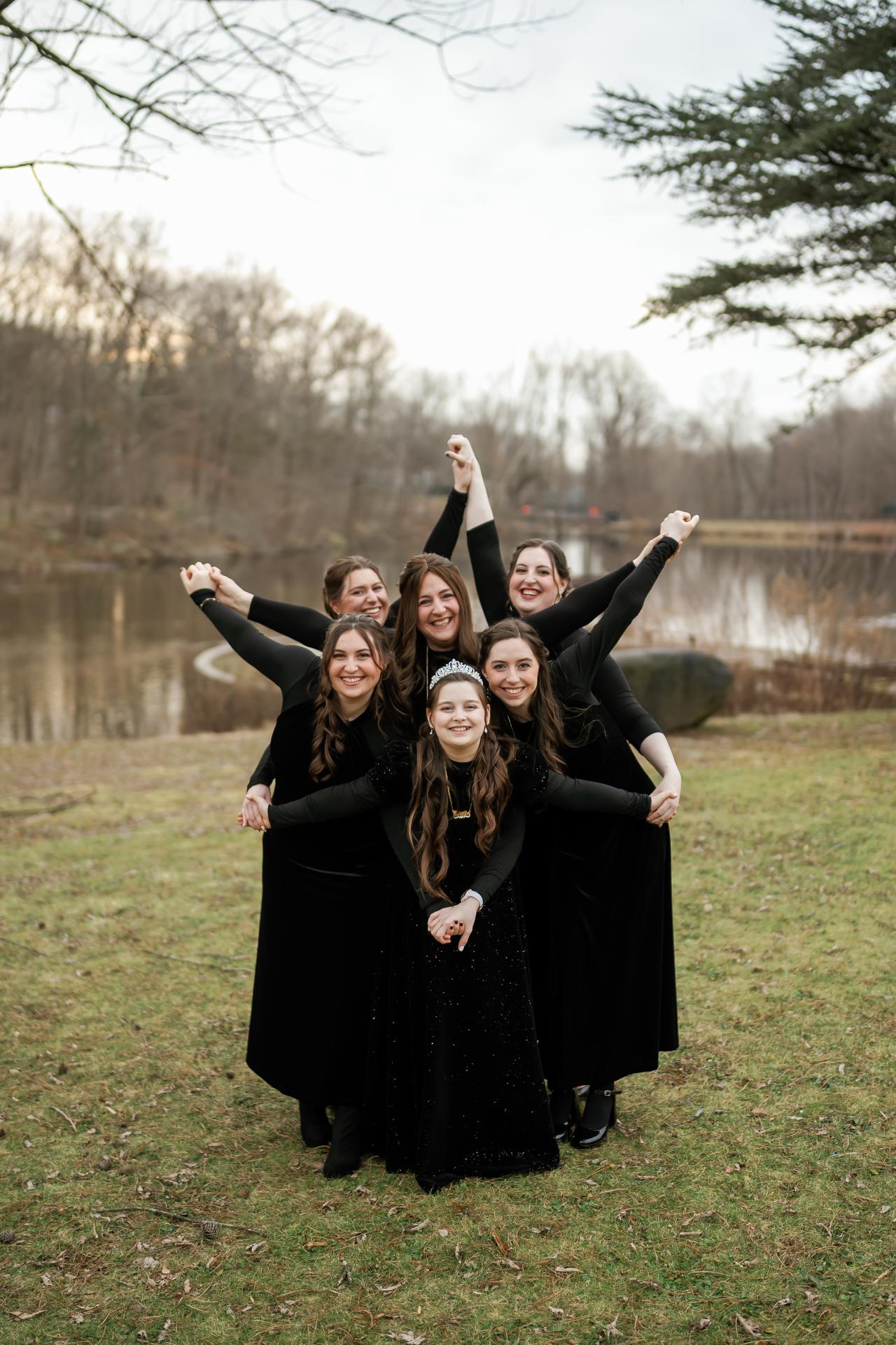 Five women in black outfits huddled together outdoors with arms raised in a joyful group hug on a grassy lawn.
