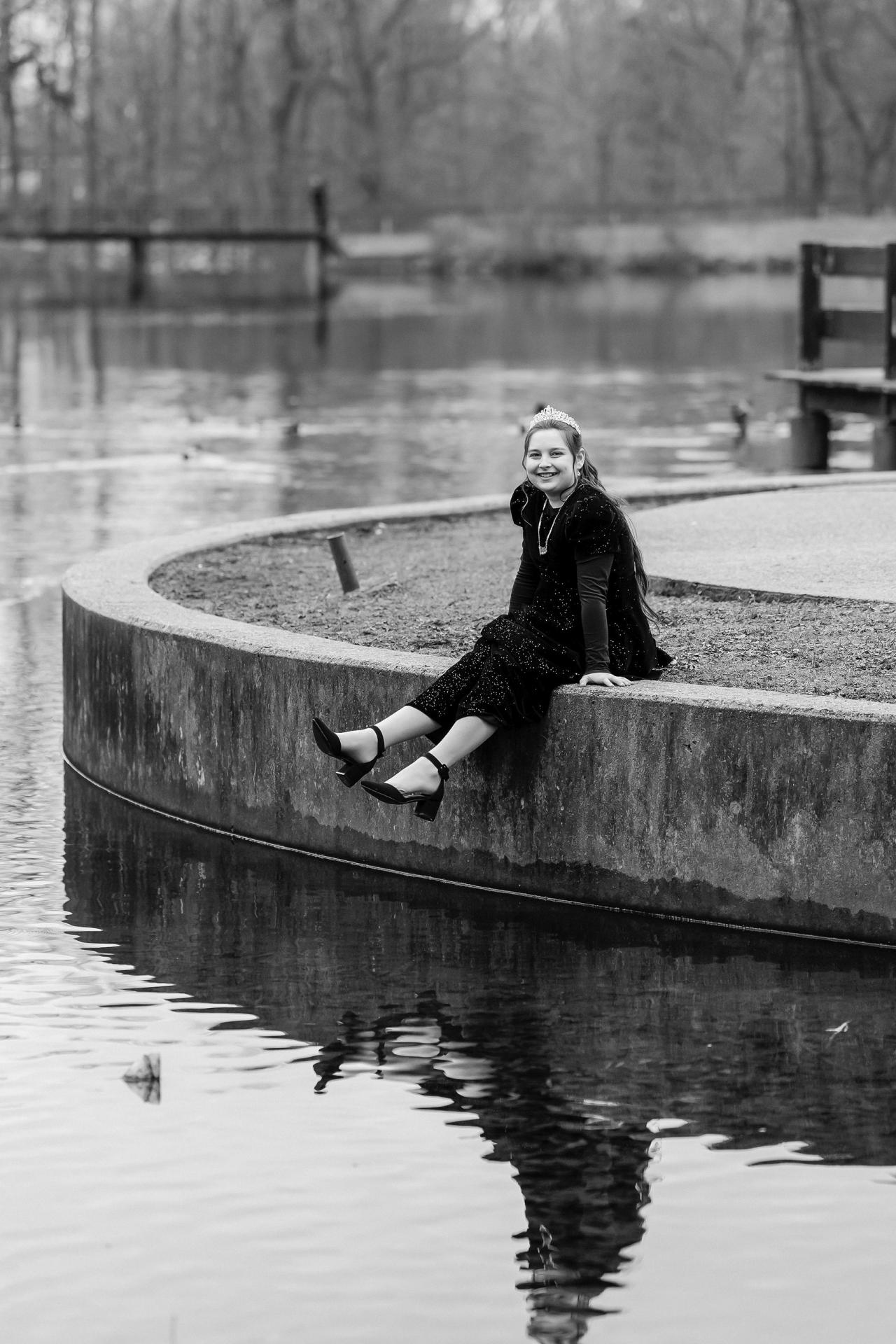 Black-and-white portrait of a smiling child seated on a stone ledge by a pond with reflective water and blurred trees in the background.