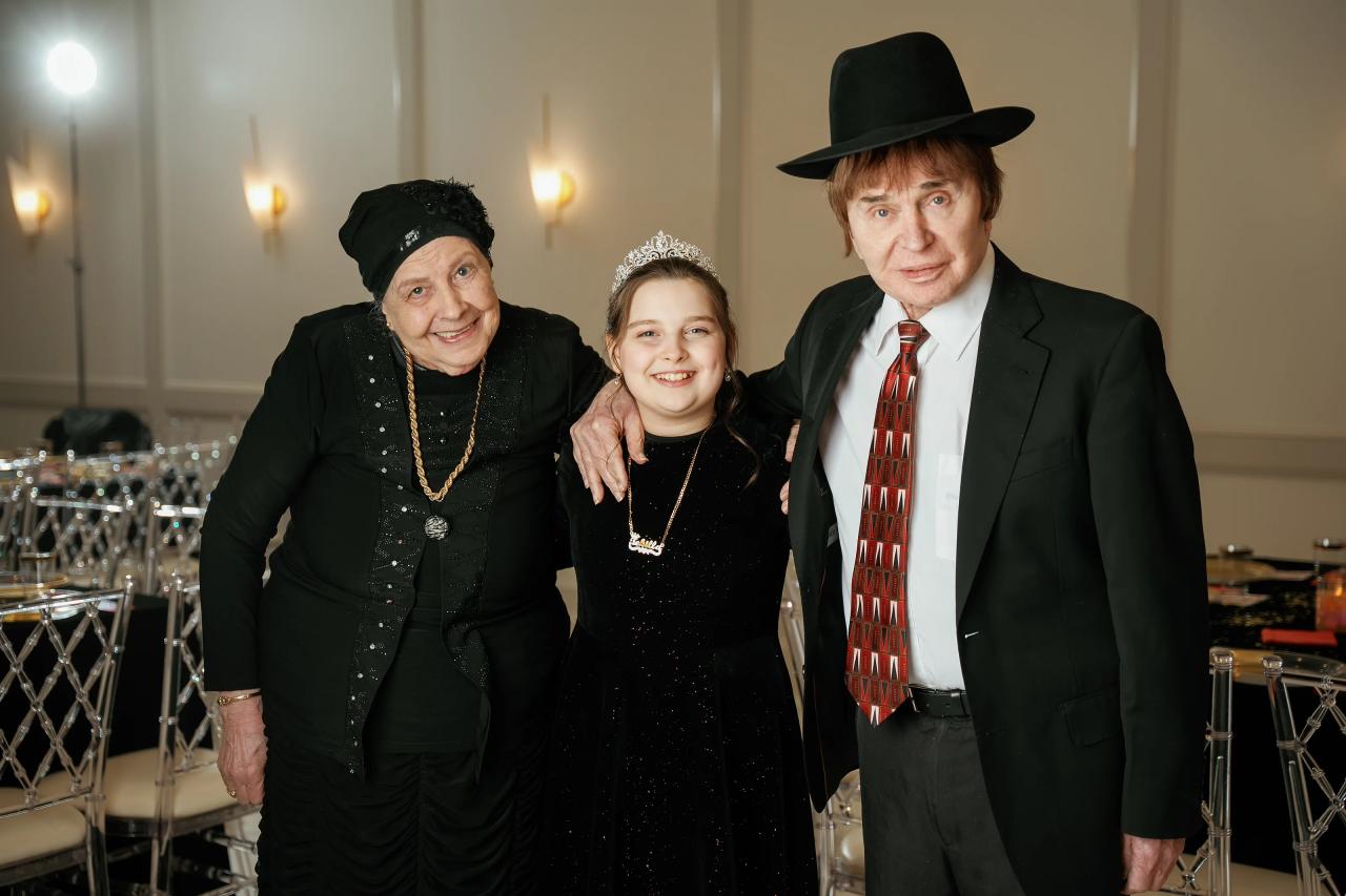 Three people in formal attire pose together indoors, smiling toward the camera at a celebration reception.