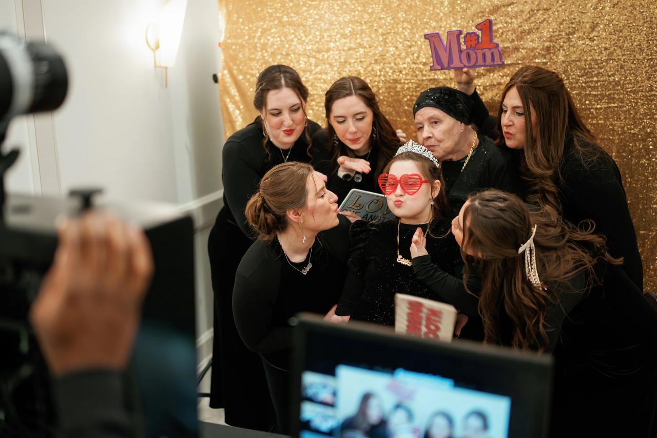 Group of women and children gathered at a “Mom #1” photo moment with a child wearing red novelty glasses against a gold sequin backdrop.