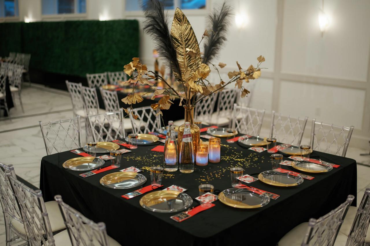 Set reception table with black tablecloth, gold feather centerpiece, candles, bottles and glassware, and red place settings in a bright indoor hall.