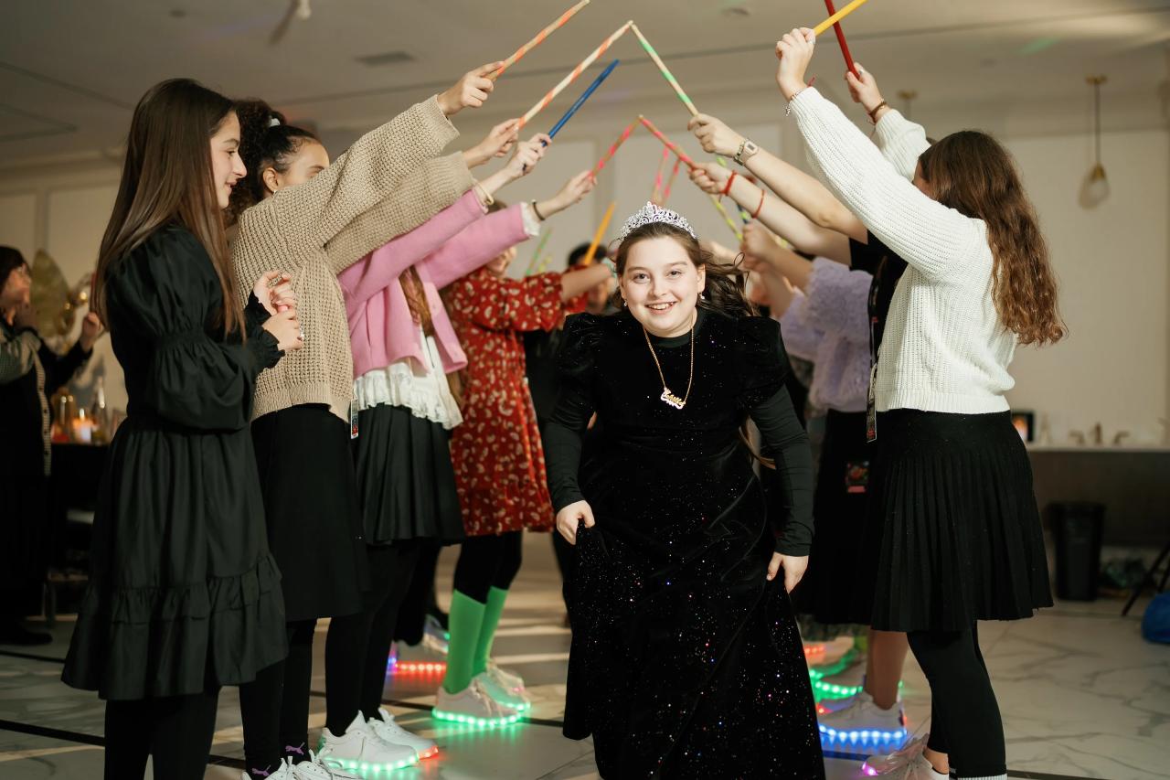 A joyful bat mitzvah girl smiling at the camera while guests surround her holding colorful ribbon streamers during a dance moment.
