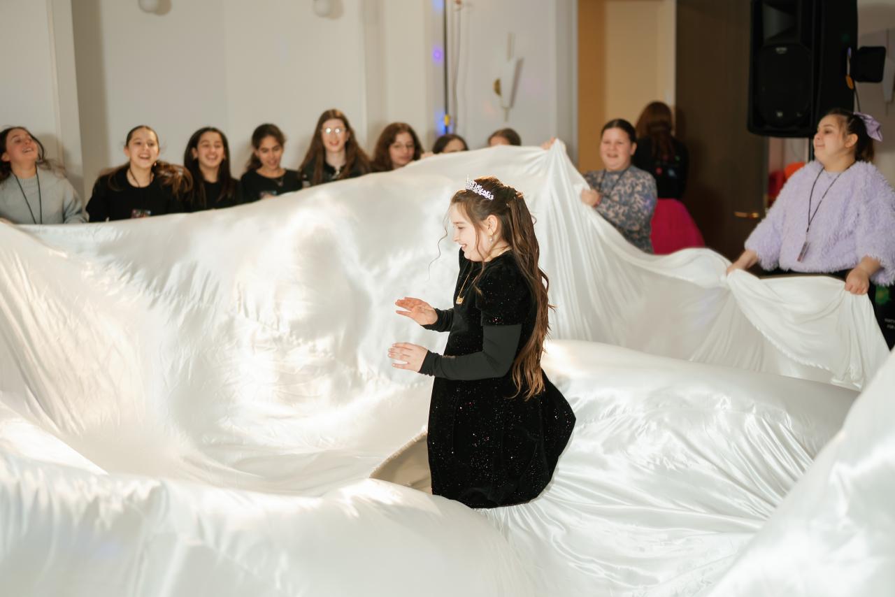 A young girl in a dark sparkly dress stands inside a white draped dance backdrop while guests watch from behind.