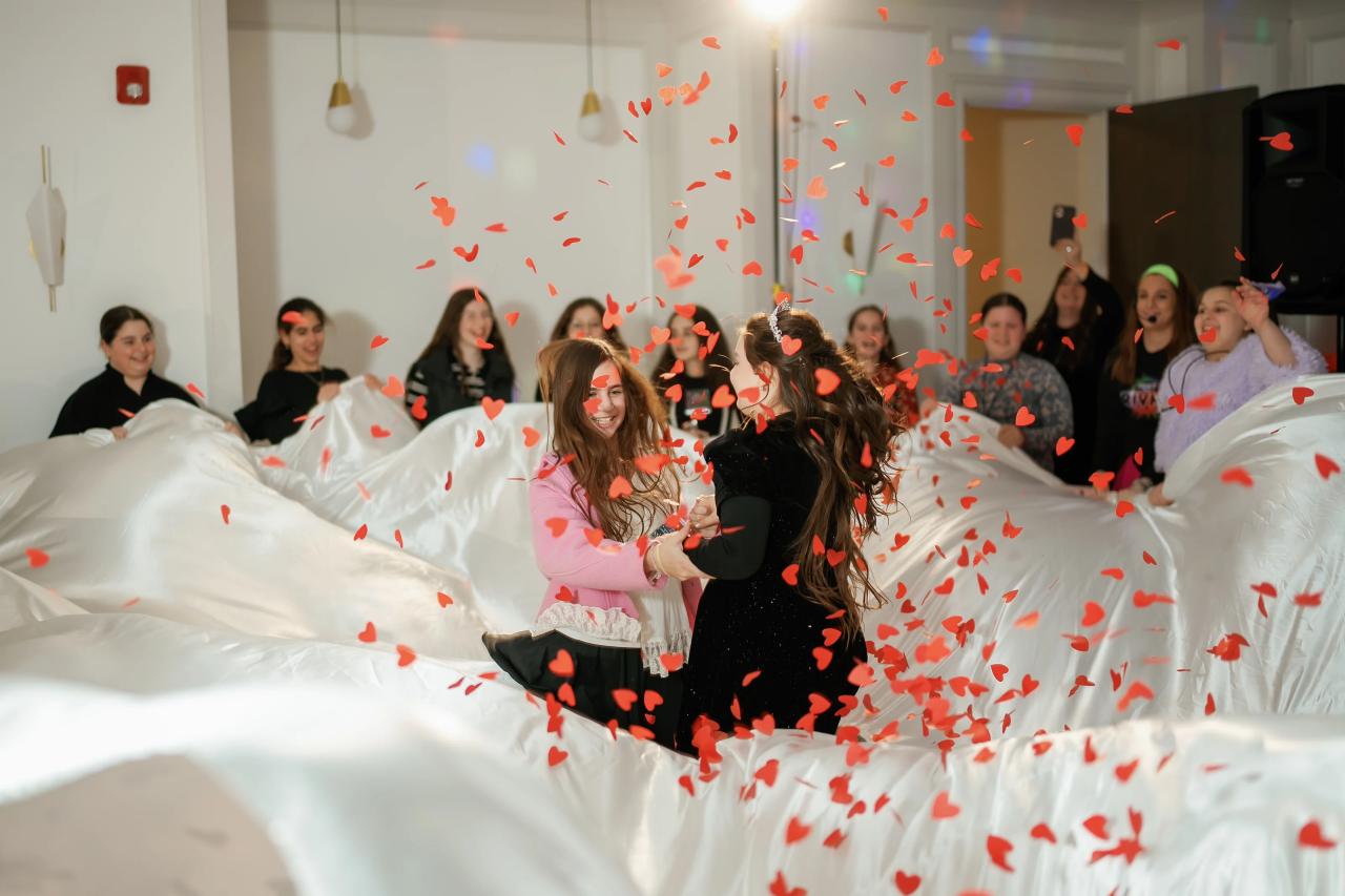 Children smiling while dancing in a hall as red confetti falls across a white-draped foreground area.