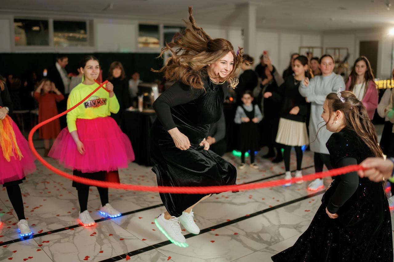 Woman in a black dress mid-dance, hair flying, smiling as she bends forward on a reception dance floor with red ribbon lines and confetti-like red pieces; guests watch in the background.