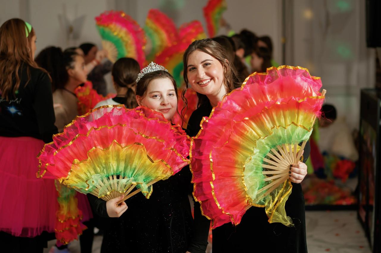 Two girls smiling and holding bright multi-colored decorative fans at a lively event.