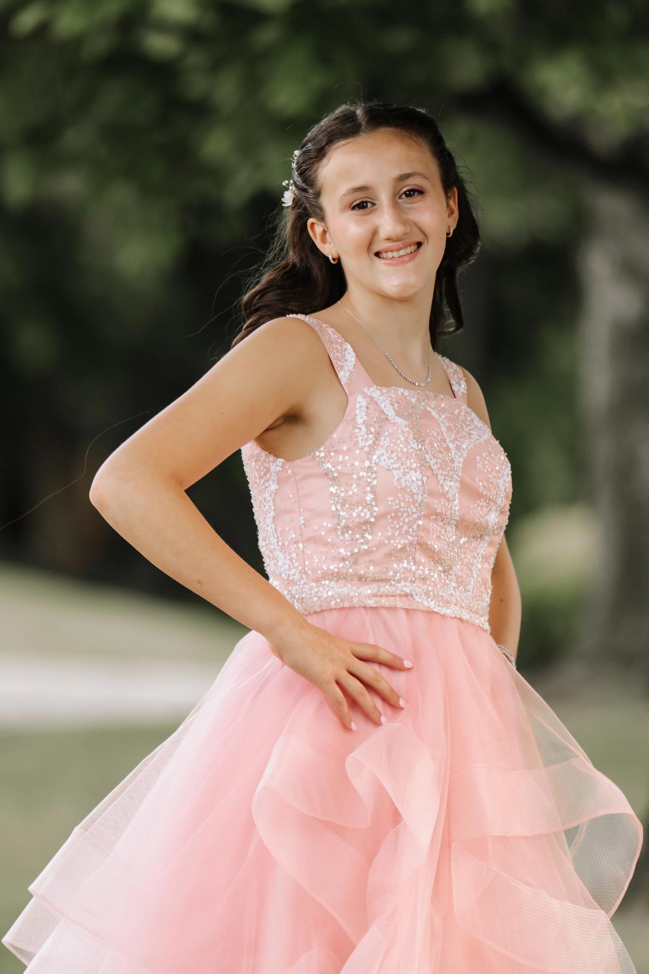 Outdoor portrait of a girl in a pink, sequined dress smiling and posing with one hand on her hip, with a softly blurred green background. Bat Mitzvah photography at Doubletree By Hilton Hotel Fort Lee - George Washington Bridge, Fort Lee, NJ.