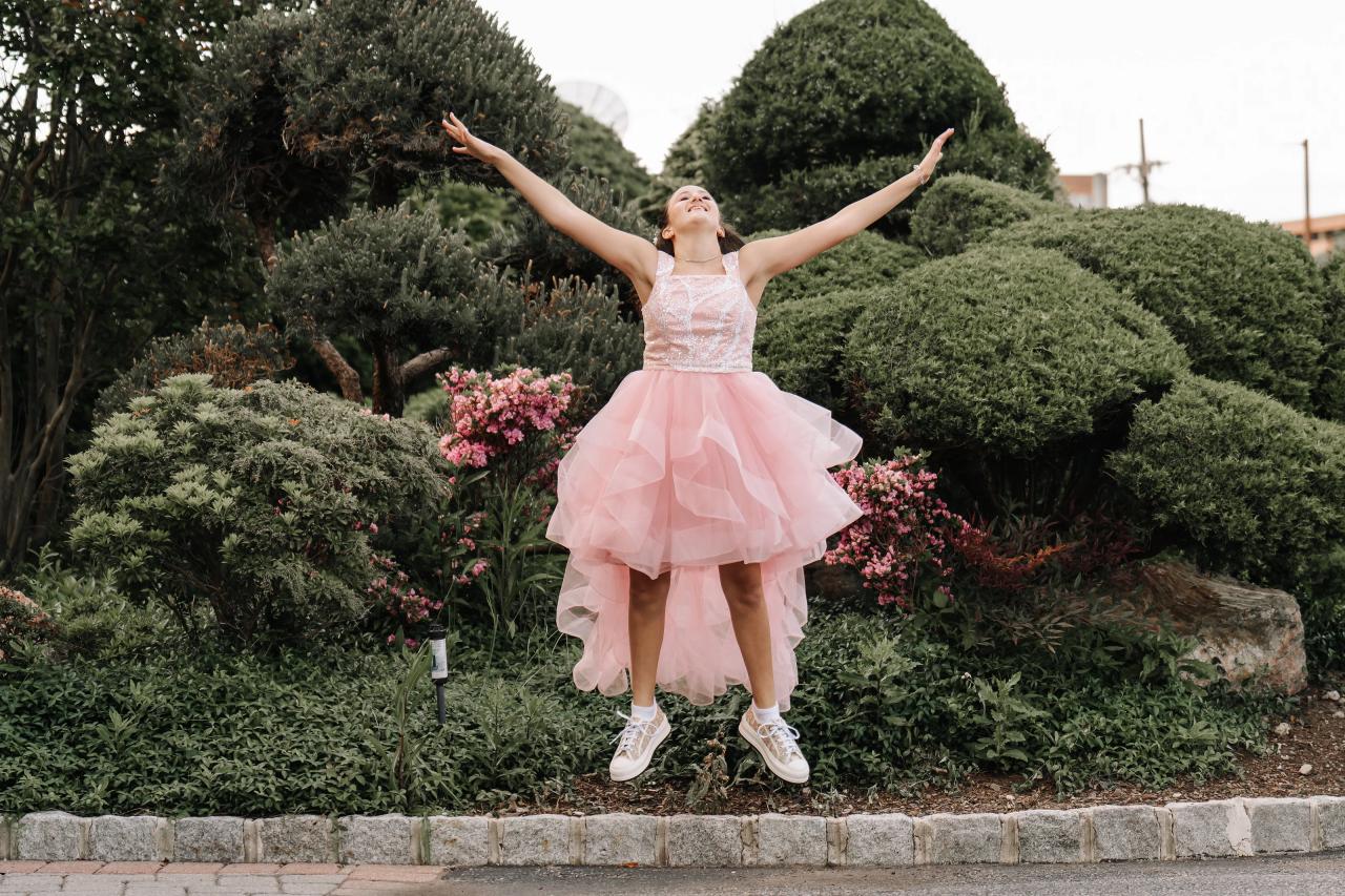 A young girl in a light pink dress stands outdoors with arms raised, smiling, surrounded by manicured hedges and pink flowers. Bat Mitzvah photography at Doubletree By Hilton Hotel Fort Lee - George Washington Bridge, Fort Lee, NJ.