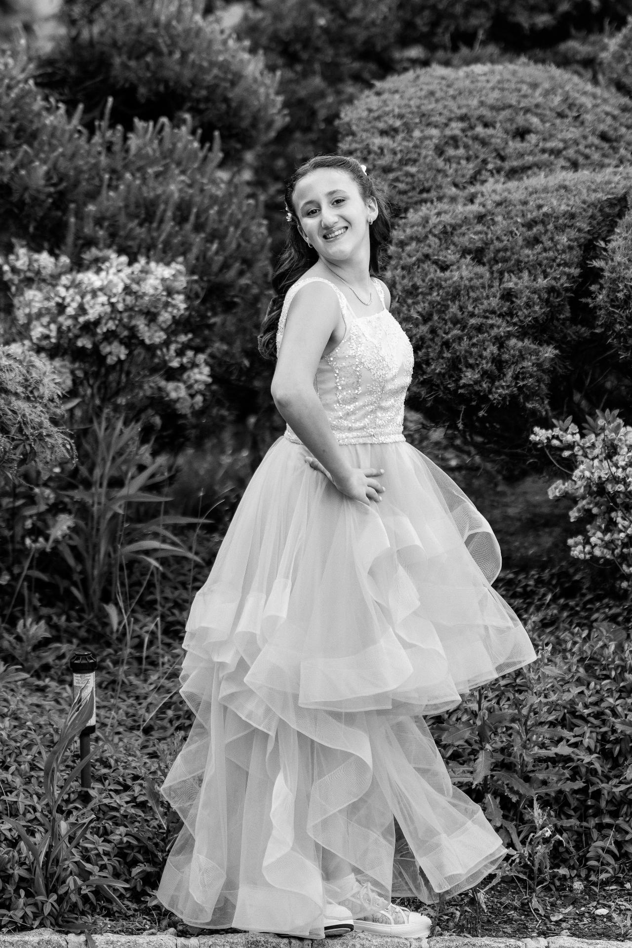 Black-and-white portrait of a young bat mitzvah girl standing in a full dress outdoors, smiling with greenery in the background. Bat Mitzvah photography at Doubletree By Hilton Hotel Fort Lee - George Washington Bridge, Fort Lee, NJ.