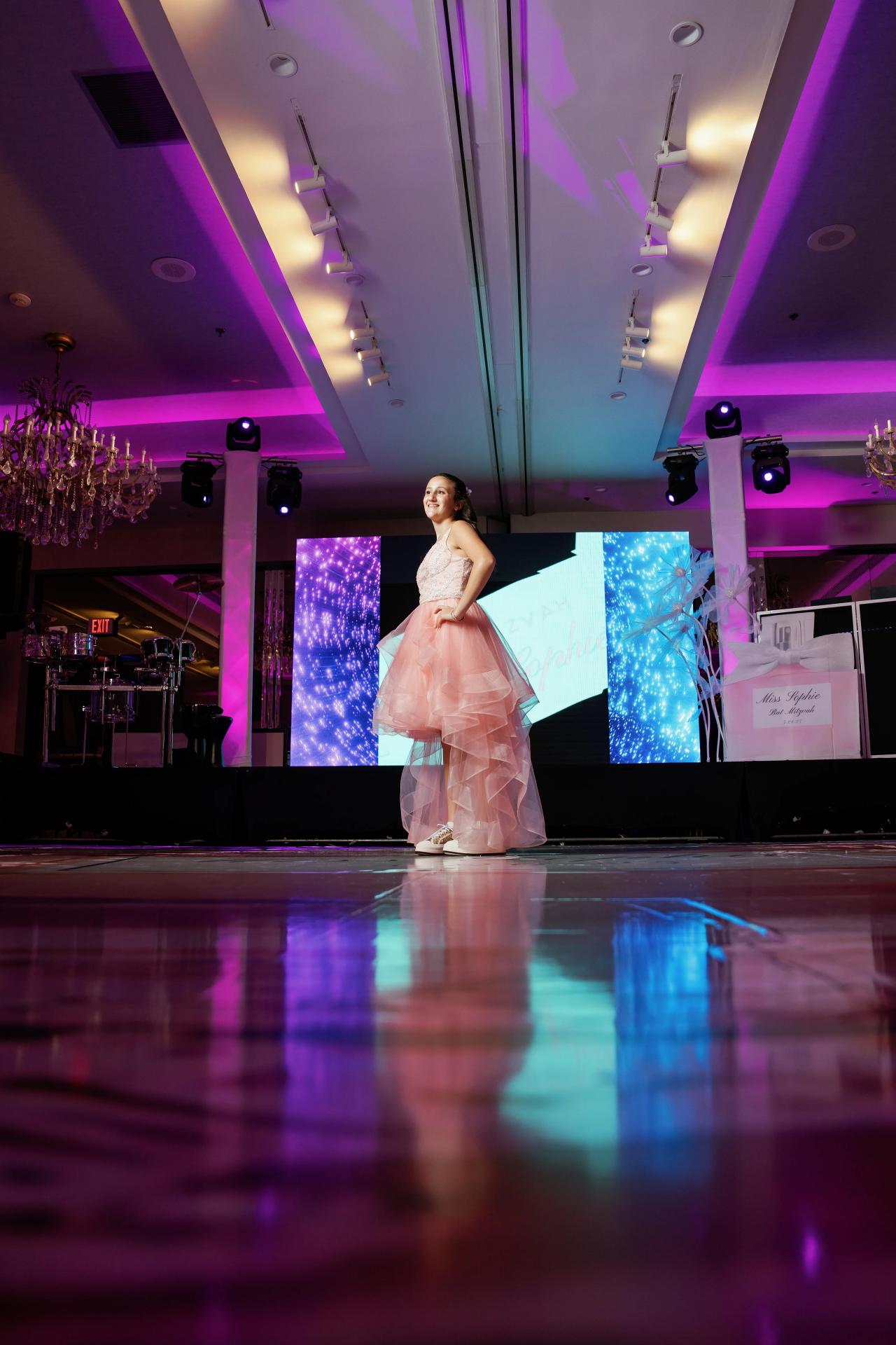 A young girl in a light-colored formal dress stands and walks on a stage in a ballroom with dramatic purple and blue event lighting and reflective floor. Bat Mitzvah photography at Doubletree By Hilton Hotel Fort Lee - George Washington Bridge, Fort Lee, NJ.