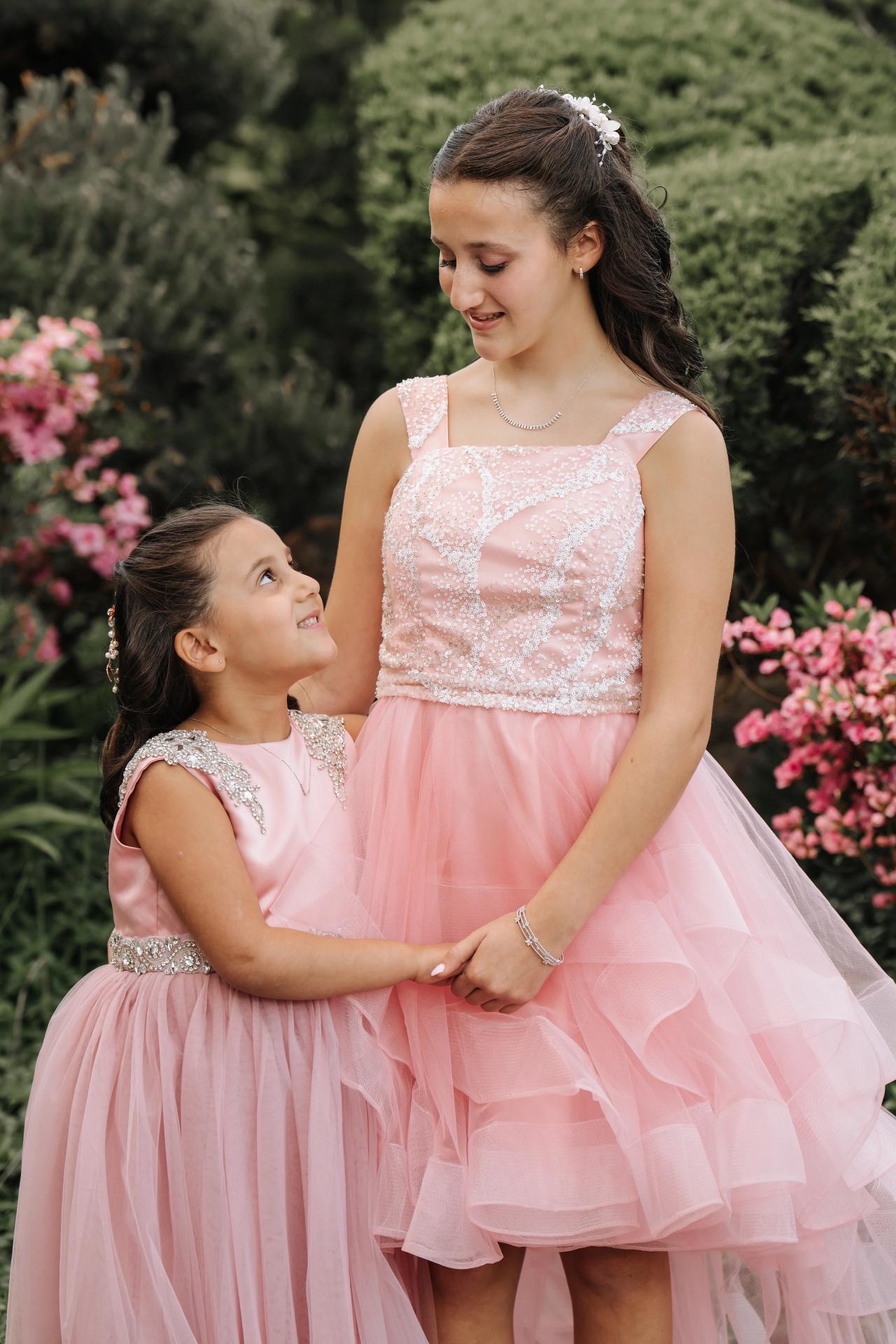 Outdoor portrait of a woman in a pink dress holding hands with a smiling young girl in a matching pink dress, surrounded by greenery and pink flowers. Bat Mitzvah photography at Doubletree By Hilton Hotel Fort Lee - George Washington Bridge, Fort Lee, NJ.