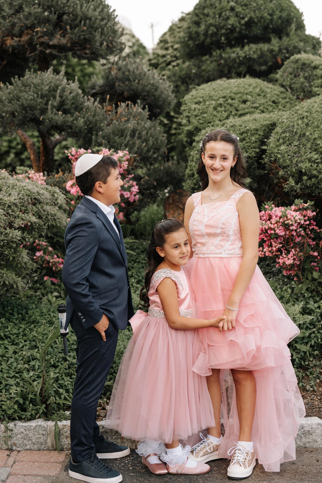 Three children dressed in formal outfits posed outdoors in a garden of hedges and pink flowers; one child smiles facing the camera while the others stand close together. Bat Mitzvah photography at Doubletree By Hilton Hotel Fort Lee - George Washington Bridge, Fort Lee, NJ.