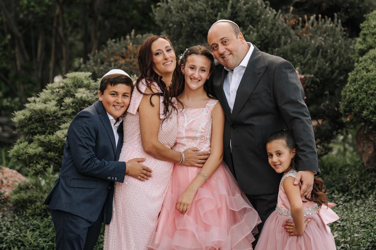 Outdoor garden family portrait of a smiling bride in a pink dress, standing with the groom and two children in pink outfits, surrounded by greenery. Bat Mitzvah photography at Doubletree By Hilton Hotel Fort Lee - George Washington Bridge, Fort Lee, NJ.