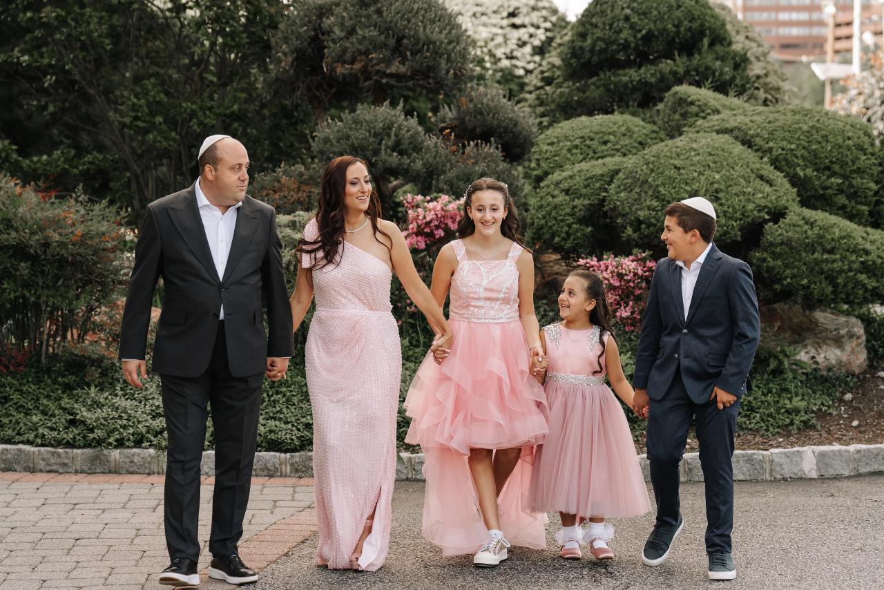 A posed family group walking hand-in-hand outdoors, with the adults and three children in coordinated attire in front of manicured hedges and pink flowers. Bat Mitzvah photography at Doubletree By Hilton Hotel Fort Lee - George Washington Bridge, Fort Lee, NJ.