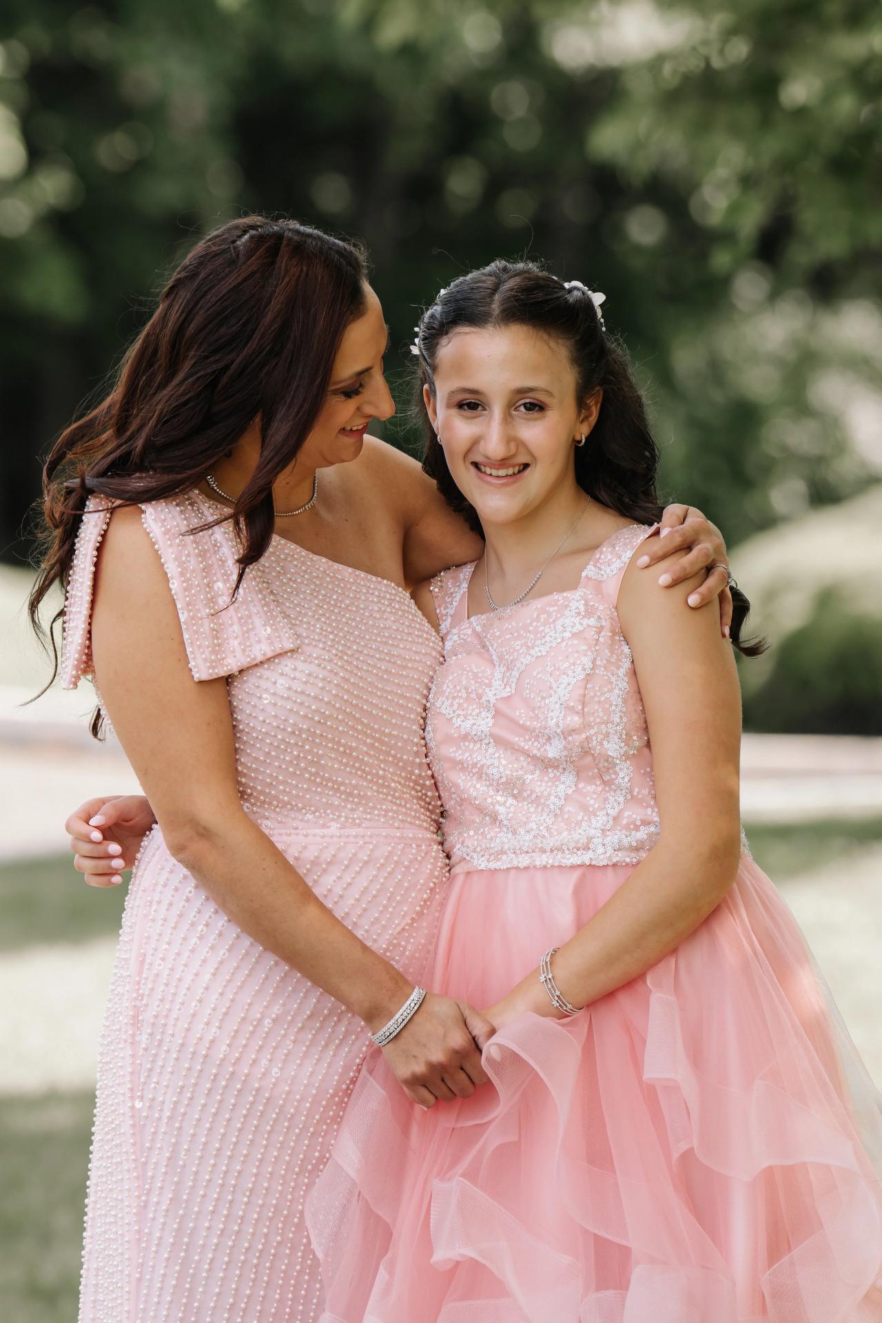 Outdoor portrait of a woman in a light pink formal dress hugging a young girl in a pink dress, both smiling, with green trees softly blurred in the background. Bat Mitzvah photography at Doubletree By Hilton Hotel Fort Lee - George Washington Bridge, Fort Lee, NJ.