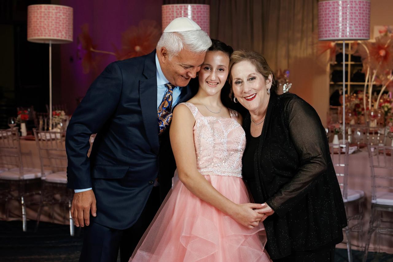 Warm reception portrait of three people posing closely together: an older man and woman on either side of a smiling girl in a pink dress, with soft event lighting and draped decor in the background. Bat Mitzvah photography at Doubletree By Hilton Hotel Fort Lee - George Washington Bridge, Fort Lee, NJ.