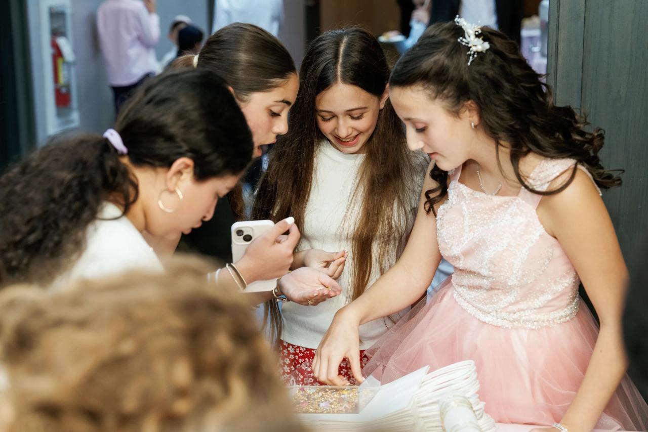 Three young girls laughing together at a crowded event table while one holds a phone and another reaches toward an open book or snack on the table. Bat Mitzvah photography at Doubletree By Hilton Hotel Fort Lee - George Washington Bridge, Fort Lee, NJ.