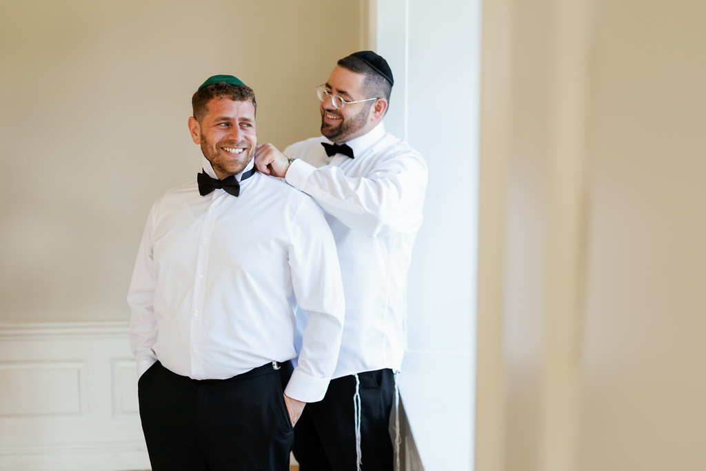Two men in black yarmulkes and white shirts share a warm smile; one adjusts the other’s clothing near the collar in a bright, minimalist indoor setting.