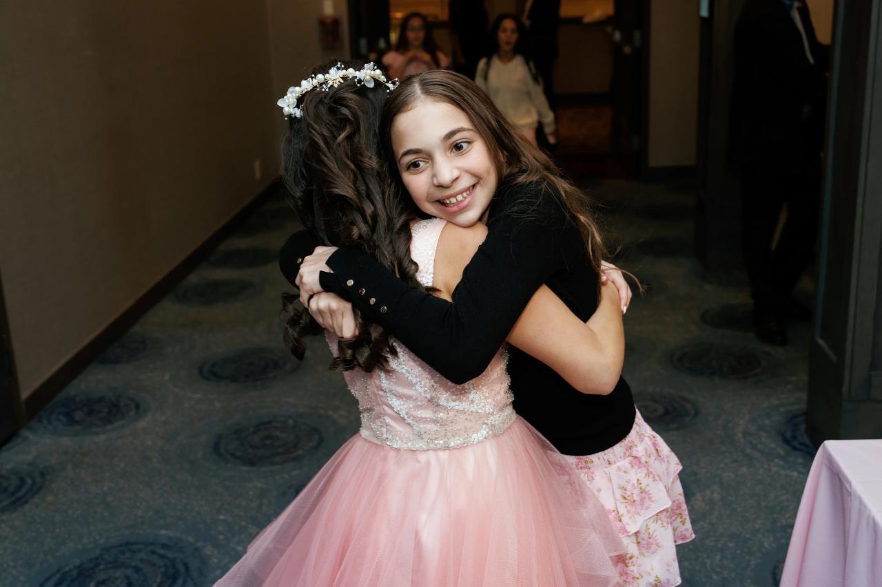 A young girl in a light pink dress is hugged and cheek-kissed by another girl in dark clothing during a wedding reception, smiling in an indoor event space. Bat Mitzvah photography at Doubletree By Hilton Hotel Fort Lee - George Washington Bridge, Fort Lee, NJ.