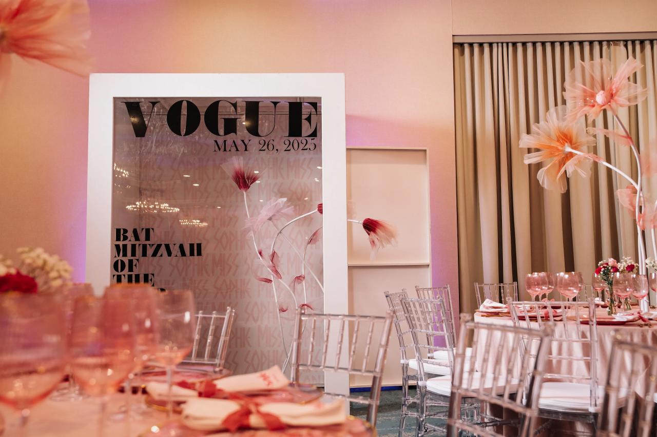 Reception table decor with a Vogue-branded magazine-style display and pink floral accents in the background, with clear chairs and place settings in the foreground. Bat Mitzvah photography at Doubletree By Hilton Hotel Fort Lee - George Washington Bridge, Fort Lee, NJ.