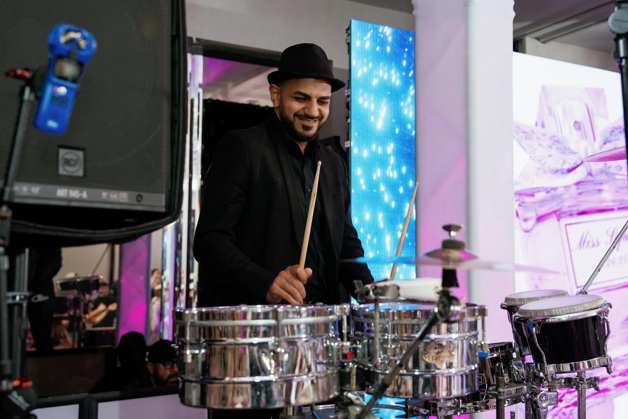 Smiling percussionist playing a drum kit on a wedding reception stage, lit by blue and purple LED lighting. Bat Mitzvah photography at Doubletree By Hilton Hotel Fort Lee - George Washington Bridge, Fort Lee, NJ.