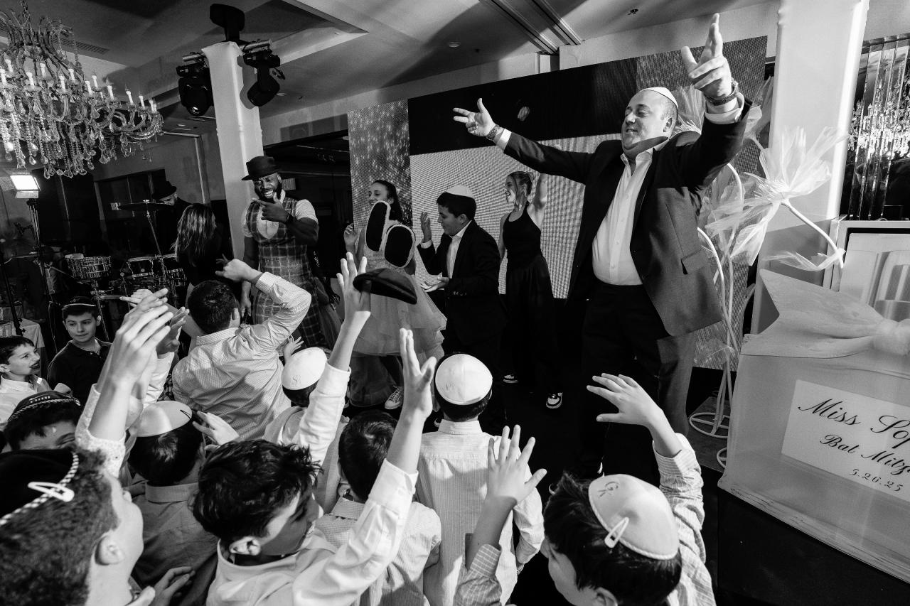 Black-and-white wedding dance-floor scene with a guest in the foreground celebrating with an arm raised as surrounding guests reach up and cheer in a crowded room. Bat Mitzvah photography at Doubletree By Hilton Hotel Fort Lee - George Washington Bridge, Fort Lee, NJ.