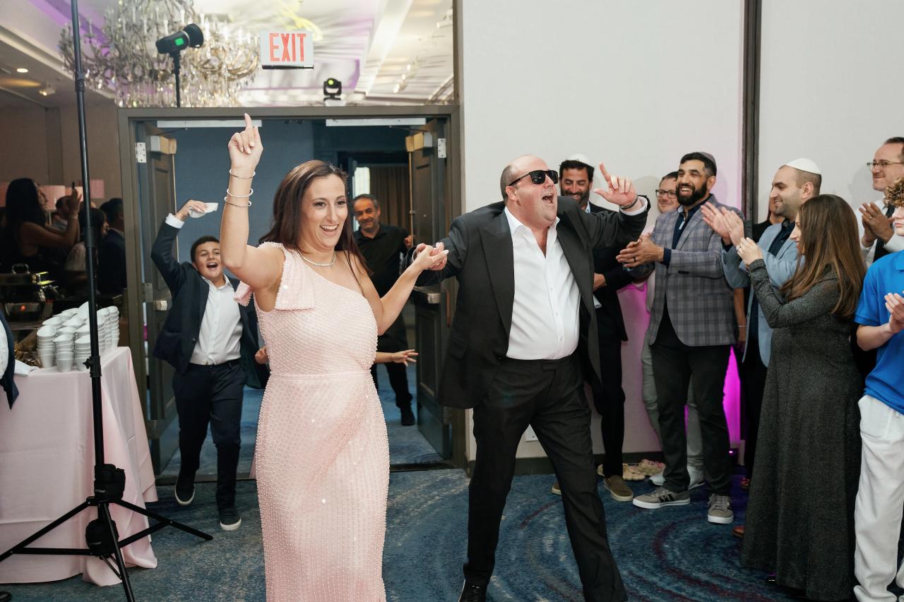 Bride in a light pink sequined dress raising her arm and smiling as the groom laughs and gestures during a lively wedding party, guests clapping along at the doorway. Bat Mitzvah photography at Doubletree By Hilton Hotel Fort Lee - George Washington Bridge, Fort Lee, NJ.