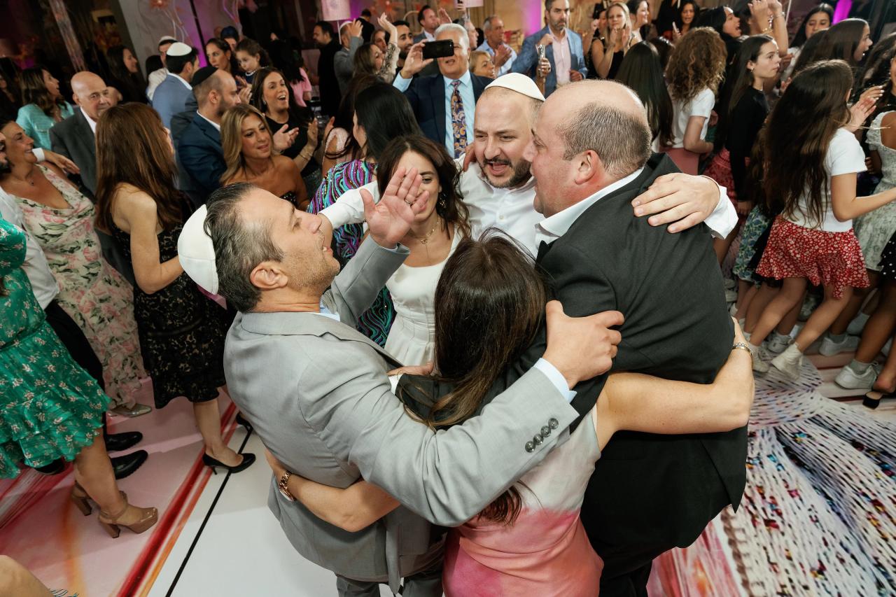 Crowded wedding dance floor with a central group hug as guests clap and cheer in colorful party lighting. Bat Mitzvah photography at Doubletree By Hilton Hotel Fort Lee - George Washington Bridge, Fort Lee, NJ.