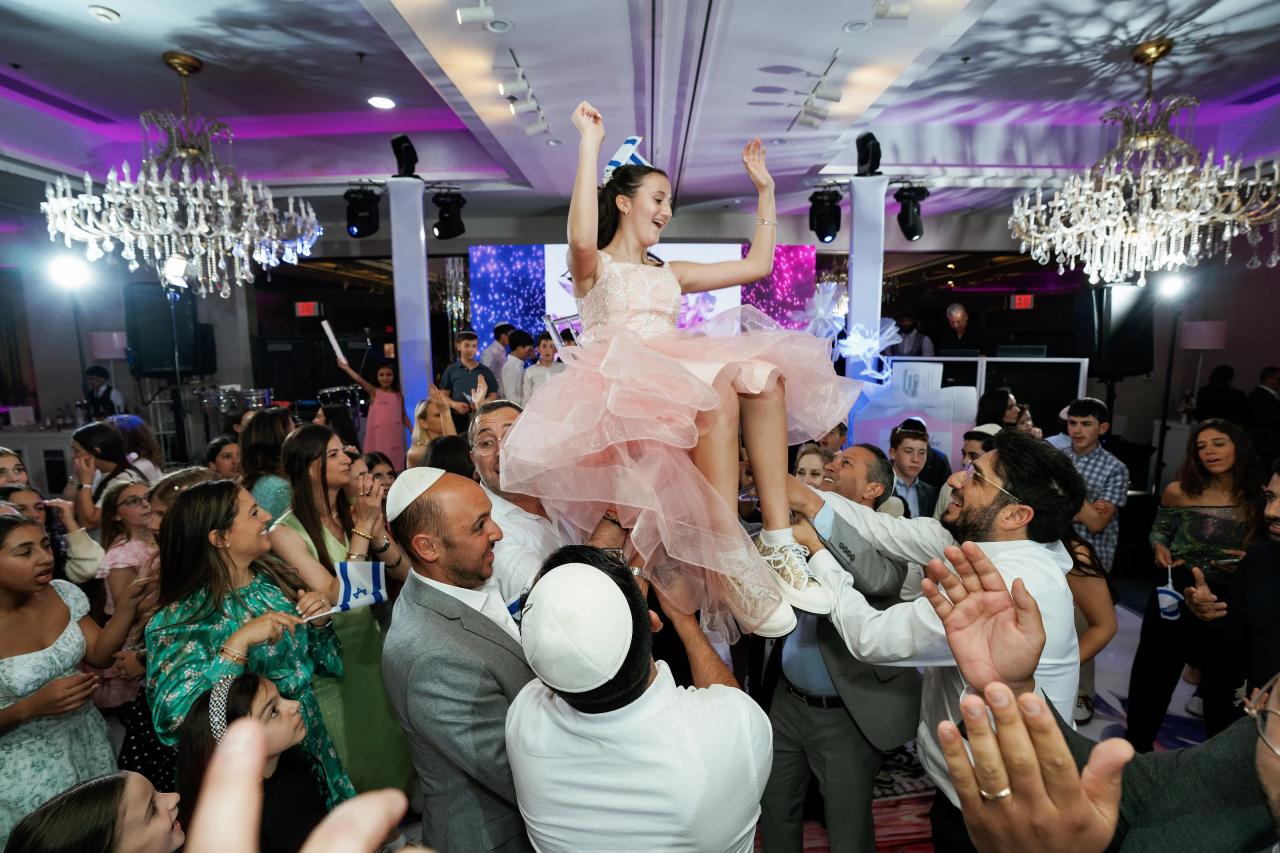 Bride in a light-colored dress leaping with both arms raised during a wedding celebration while guests crowd the dance floor reaching toward her under chandelier lighting. Bat Mitzvah photography at Doubletree By Hilton Hotel Fort Lee - George Washington Bridge, Fort Lee, NJ.