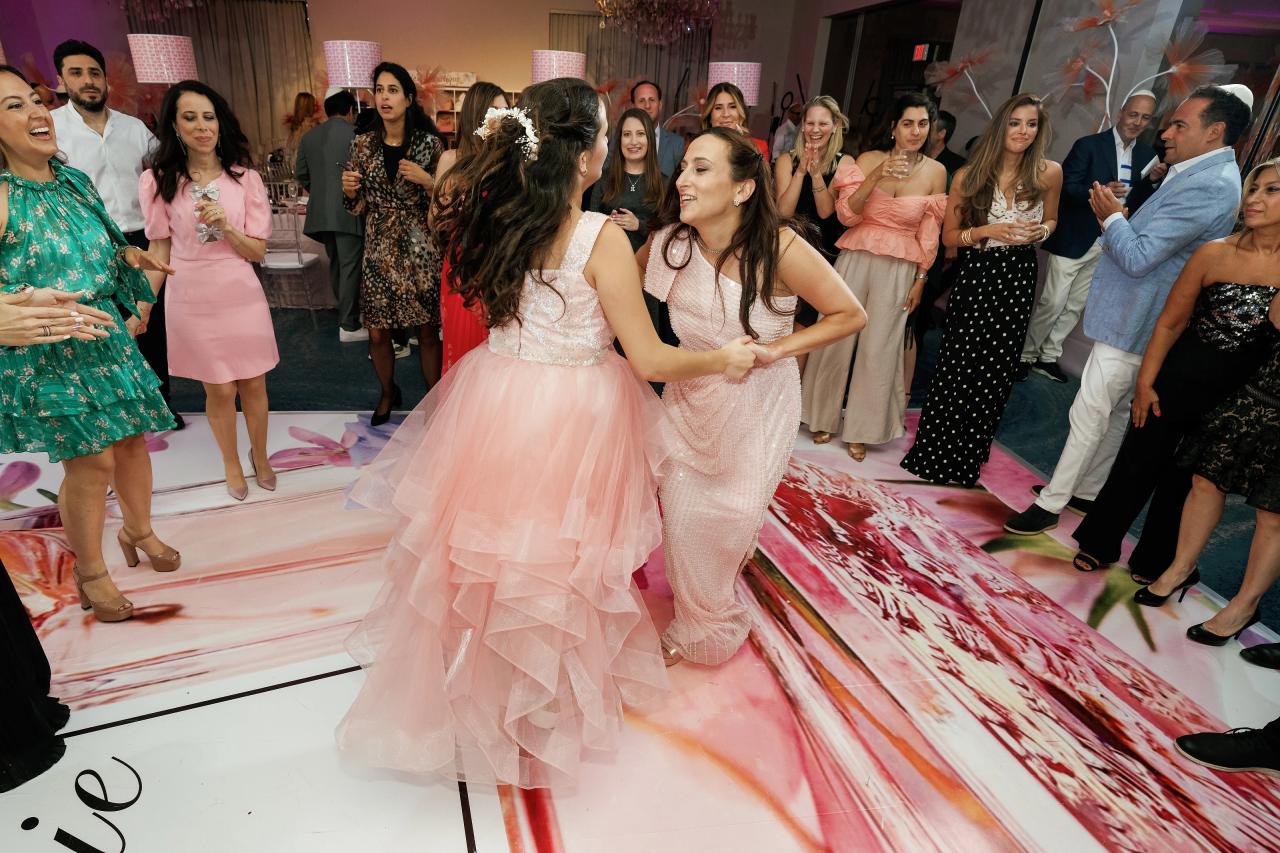 Bride and a second woman dancing and laughing on a pink-themed dance floor at a wedding reception, surrounded by guests who are clapping and watching. Bat Mitzvah photography at Doubletree By Hilton Hotel Fort Lee - George Washington Bridge, Fort Lee, NJ.