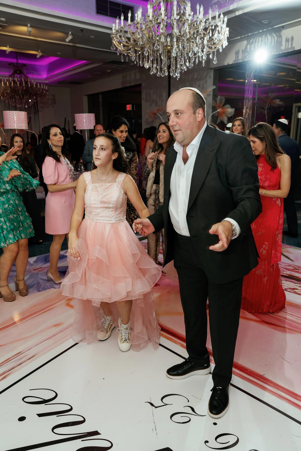 A suited man walks hand-in-hand with a young girl in a sparkly pink dress on a decorated dance floor at a celebration, with chandeliers and guests in the background. Bat Mitzvah photography at Doubletree By Hilton Hotel Fort Lee - George Washington Bridge, Fort Lee, NJ.
