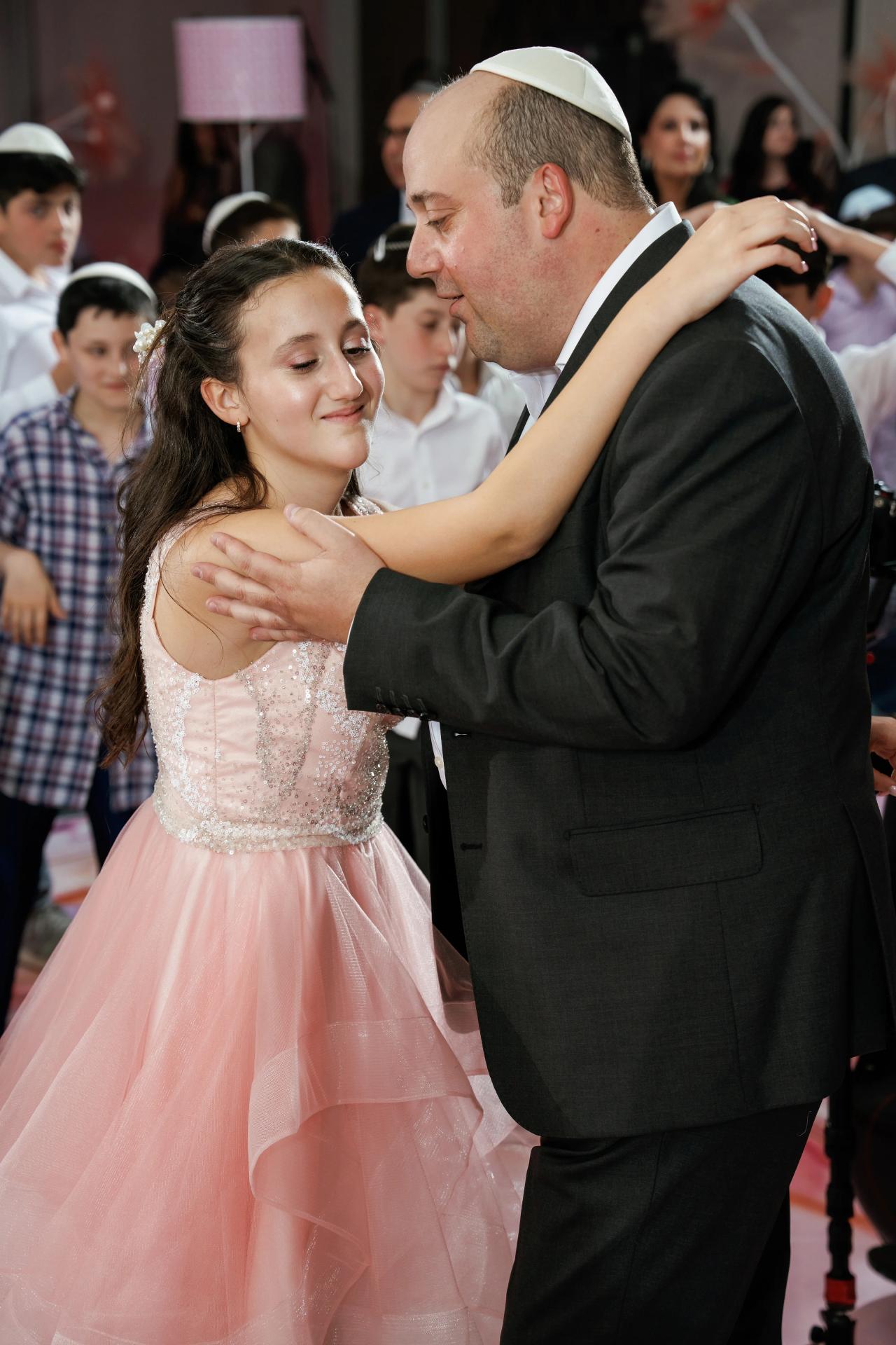 Bride in a blush dress and groom in a dark suit share an intimate dance-floor hug, eyes closed and smiling, with guests softly blurred behind them. Bat Mitzvah photography at Doubletree By Hilton Hotel Fort Lee - George Washington Bridge, Fort Lee, NJ.
