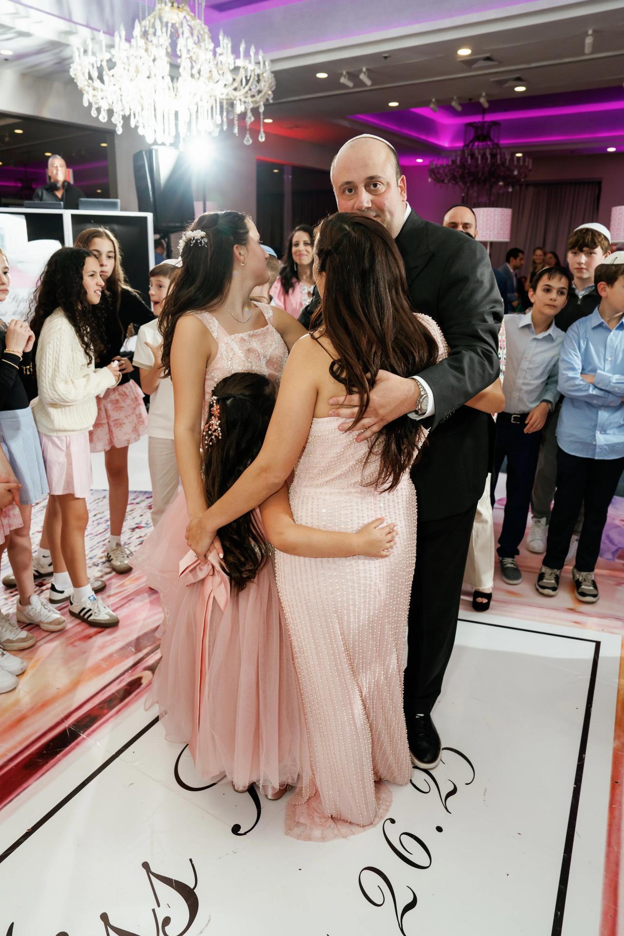 Bride in a sparkly pale gown and veil embraces with the groom on a dance floor while family and guests watch under chandelier lights and purple uplighting. Bat Mitzvah photography at Doubletree By Hilton Hotel Fort Lee - George Washington Bridge, Fort Lee, NJ.