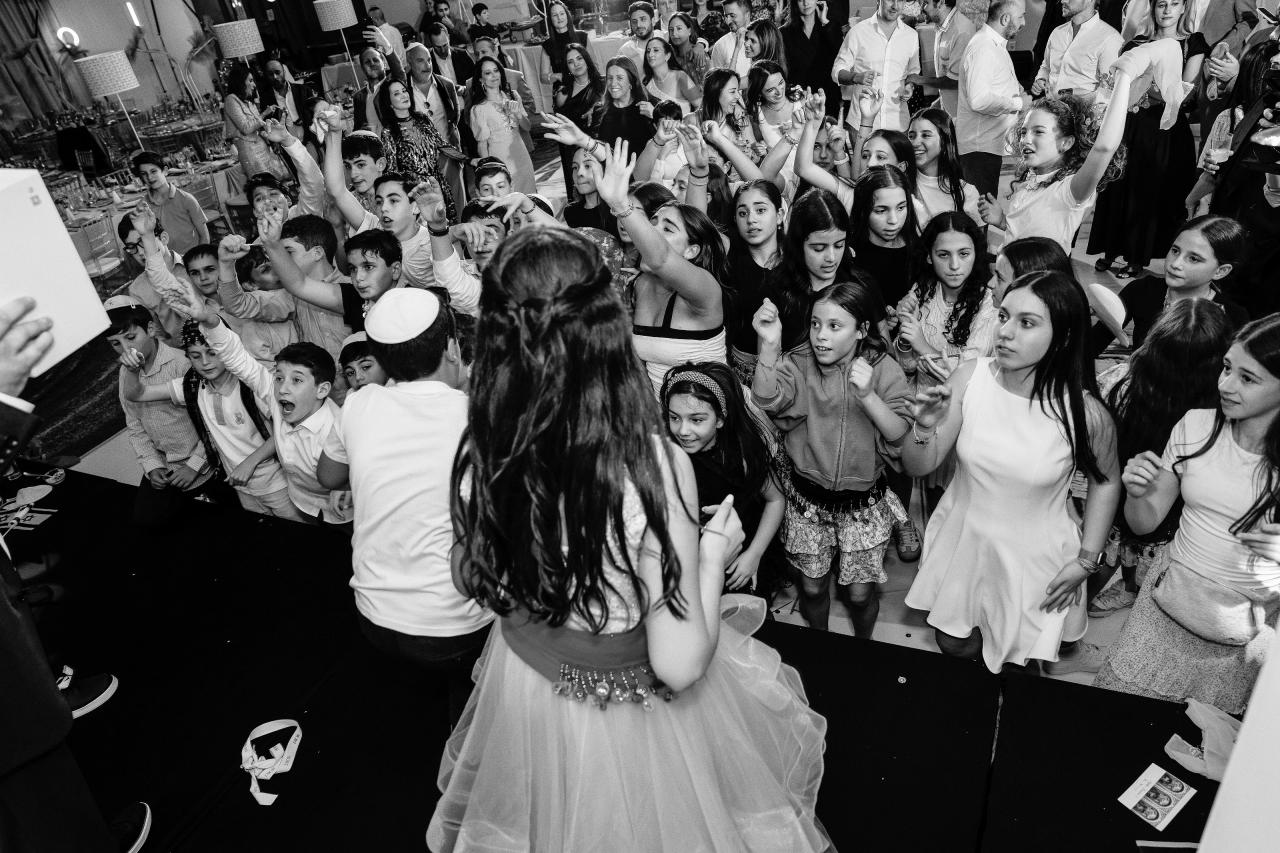 Black-and-white candid of a crowded dance floor with children and guests cheering and waving, arms raised, captured from behind the crowd. Bat Mitzvah photography at Doubletree By Hilton Hotel Fort Lee - George Washington Bridge, Fort Lee, NJ.