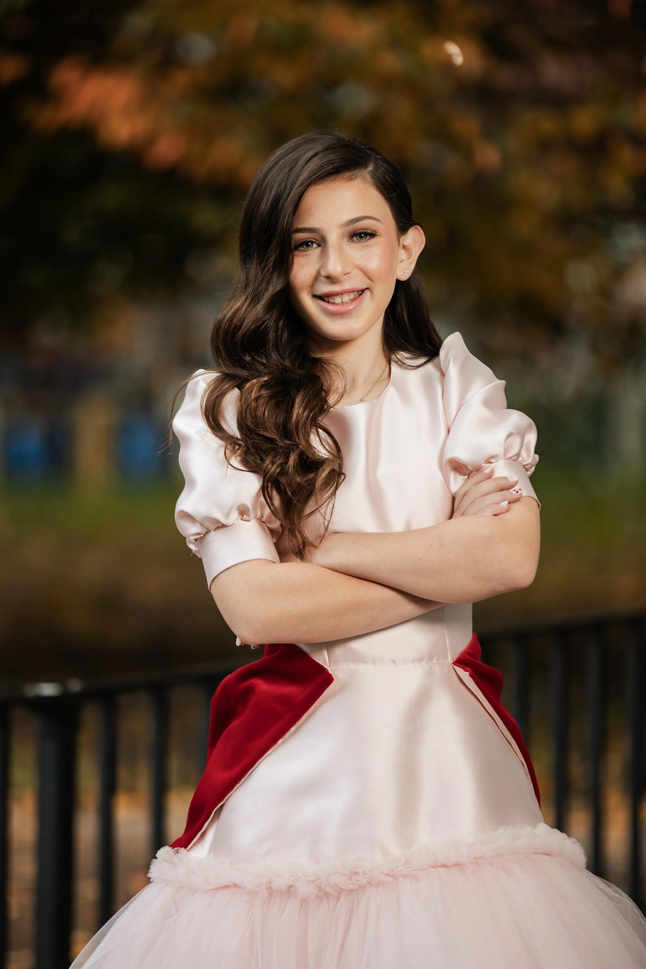 Portrait of a mitzvah girl in a white dress with red ribbon accents, arms crossed, smiling against a blurred outdoor background. Bat Mitzvah photography at Bison & Bourbon, Brooklyn, NY.