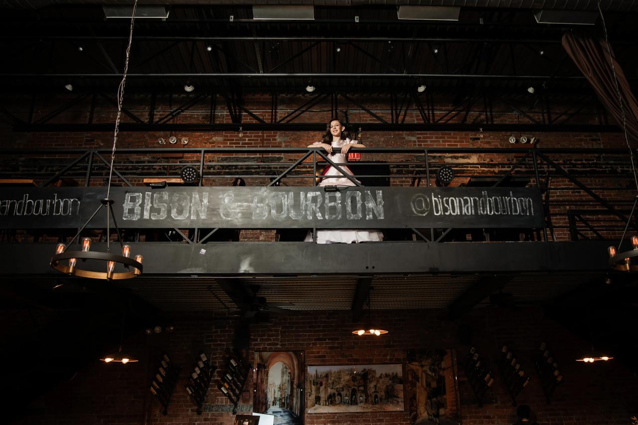 Wide shot of an indoor wedding hall showing the elevated stage area with a central person silhouetted above brick and industrial beams; large banner text spans the upper wall. Bat Mitzvah photography at Bison & Bourbon, Brooklyn, NY.