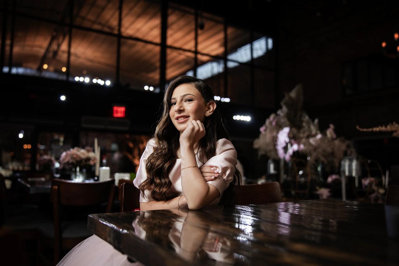 Candid event photo of a smiling young woman laughing while sitting at a reflective table, with a softly lit bar and floral decor in the background. Bat Mitzvah photography at Bison & Bourbon, Brooklyn, NY.