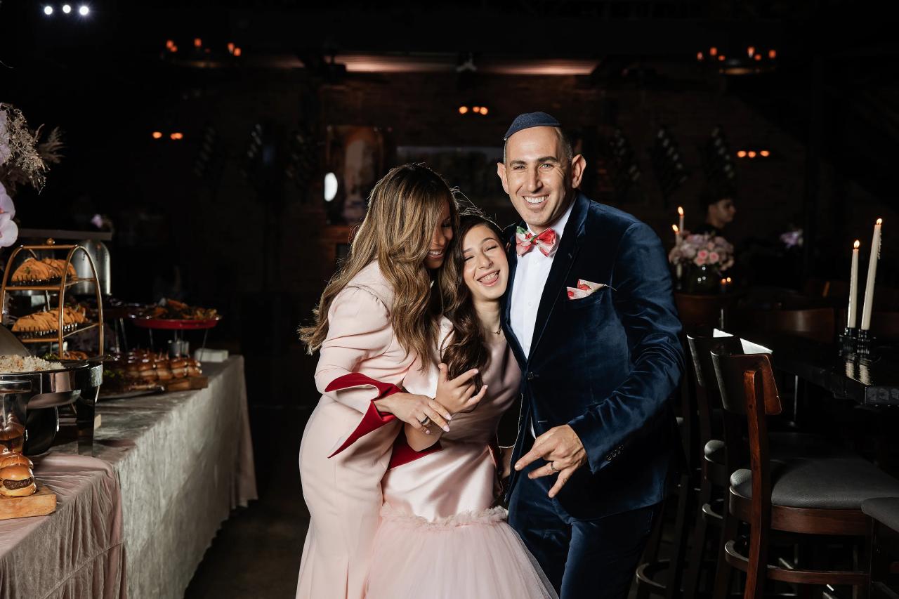 Smiling bride and groom hugging closely with a child at a warmly lit reception, candles and dining setup in the background. Bat Mitzvah photography at Bison & Bourbon, Brooklyn, NY.
