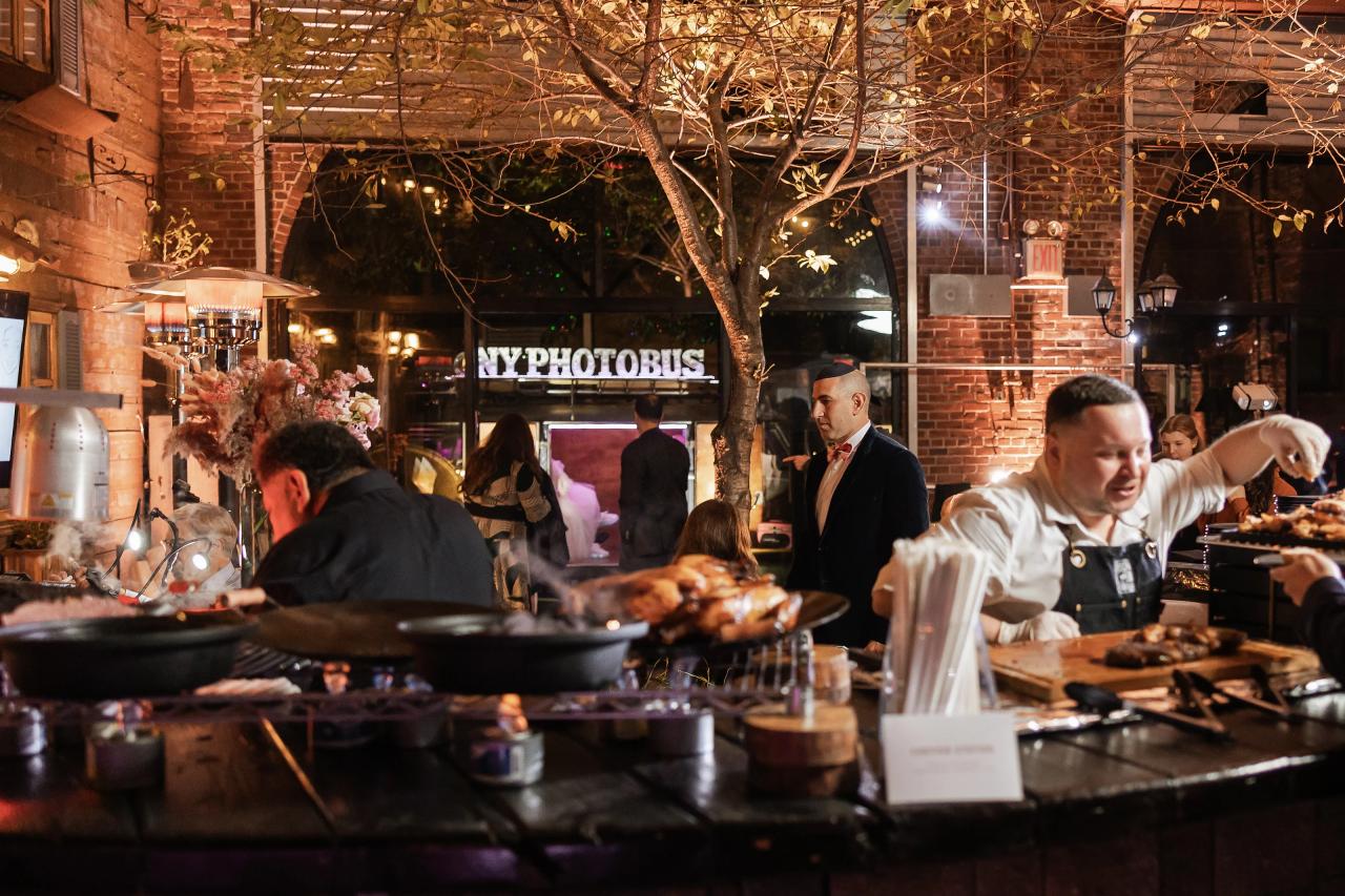 Warm indoor reception scene with a lively food station in the foreground, cookware on counters, and guests and staff gathered in the background near brick walls and lantern-style lights. Bat Mitzvah photography at Bison & Bourbon, Brooklyn, NY.