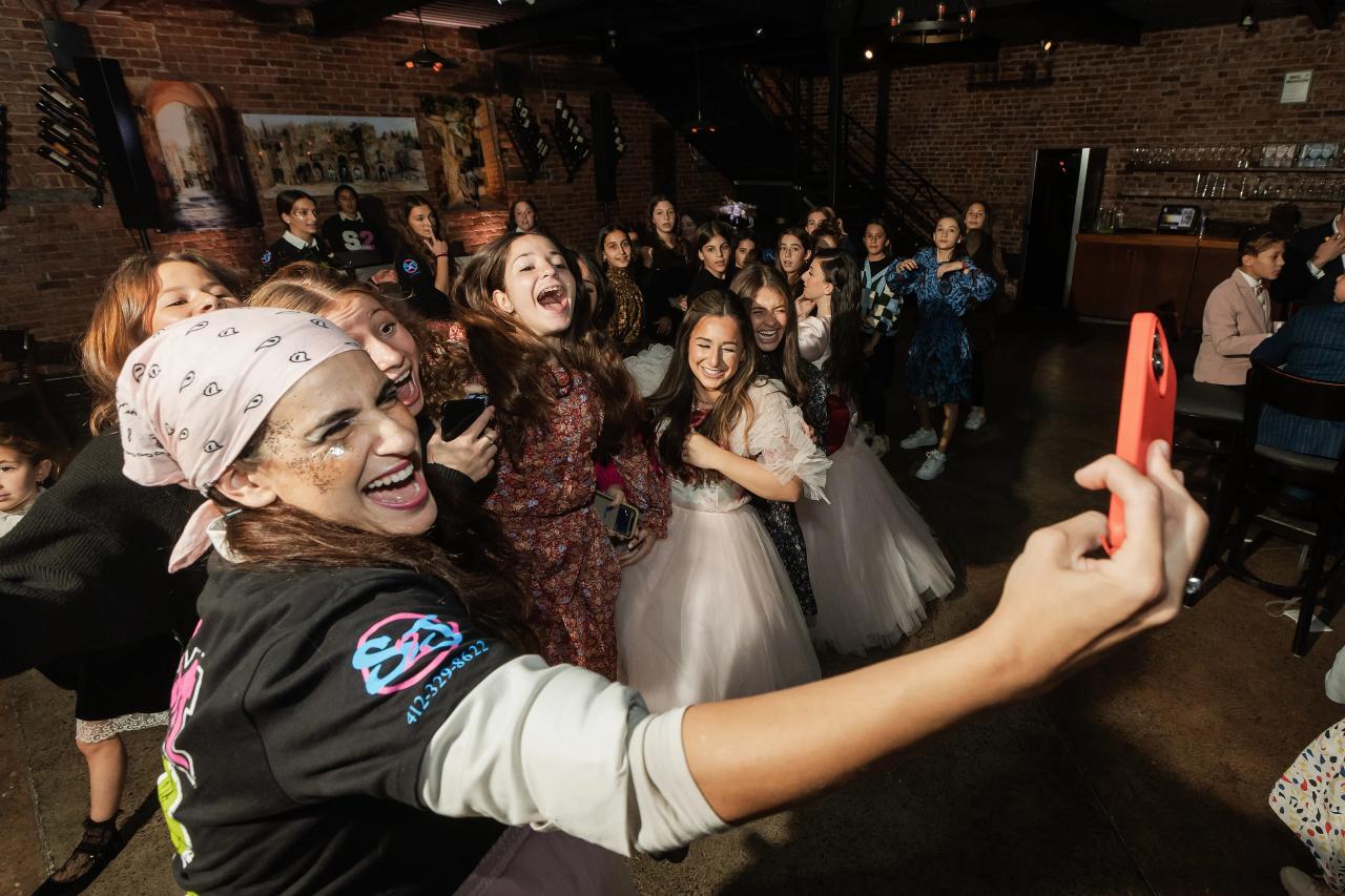 At a lively indoor simcha reception, a smiling guest in the foreground reaches toward the camera while a group laughs and dances behind her under warm low lighting. Bat Mitzvah photography at Bison & Bourbon, Brooklyn, NY.