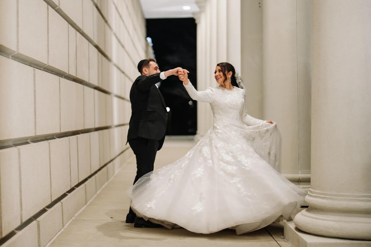 Bride in a white wedding gown and groom in a black suit dance together in a hallway, smiling as they spin in synchronized motion.