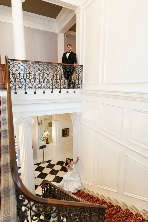 Bride standing in a white dress on a lower level while the groom in a black suit waits on the balcony above in a grand interior stairwell.