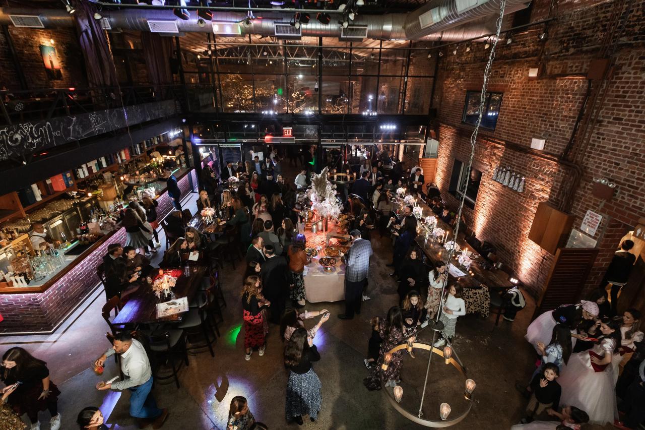 Elevated wide shot of a wedding reception ballroom with guests gathered around tables and a bar, brick walls, and colorful stage lighting. Bat Mitzvah photography at Bison & Bourbon, Brooklyn, NY.
