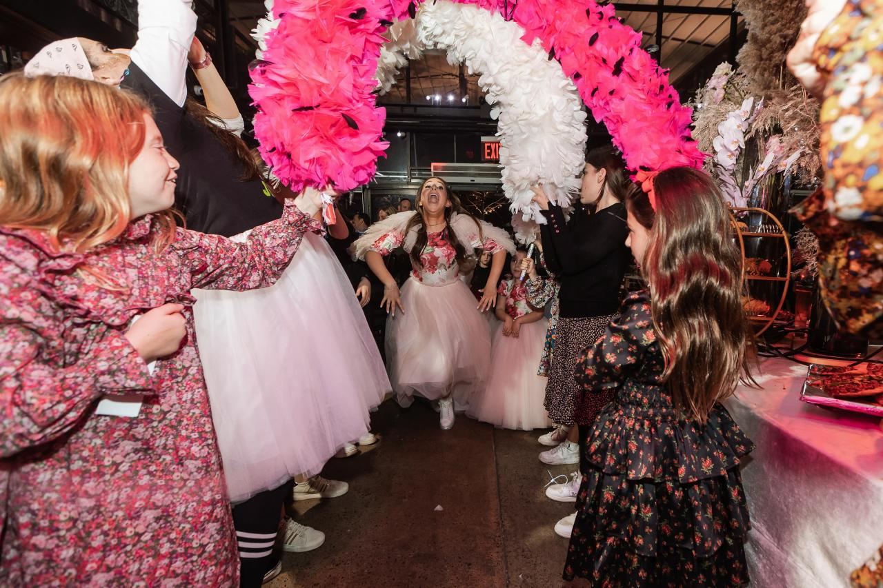 Children dancing and cheering around a girl in a light-colored dress under oversized pink-and-white decorative arches at a celebration event. Bat Mitzvah photography at Bison & Bourbon, Brooklyn, NY.