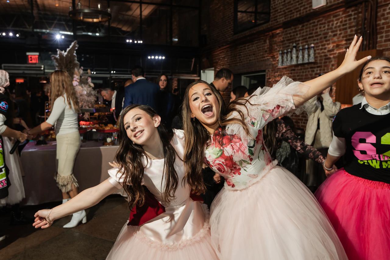 Two children dancing and laughing with arms raised at a celebration inside a brick event hall, confetti in the air and guests in the background. Bat Mitzvah photography at Bison & Bourbon, Brooklyn, NY.